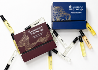 Two boxed fragrance samples and several clear glass perfume vials with black caps are arranged on a white background. The boxes and vials are branded Thousand Mornings, with one box in deep burgundy and one in dark blue, both with gold abstract line art.