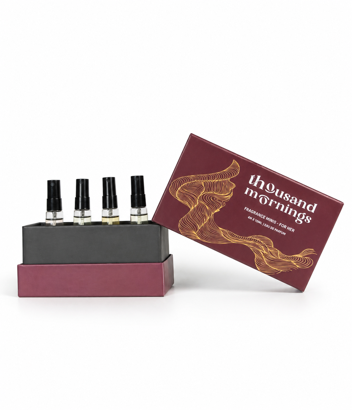 A boxed fragrance set on a white background, featuring four small spray vials in a dark tray beside an open burgundy box with gold line art and the text 'thousand mornings' and 'fragrance oil for men'.