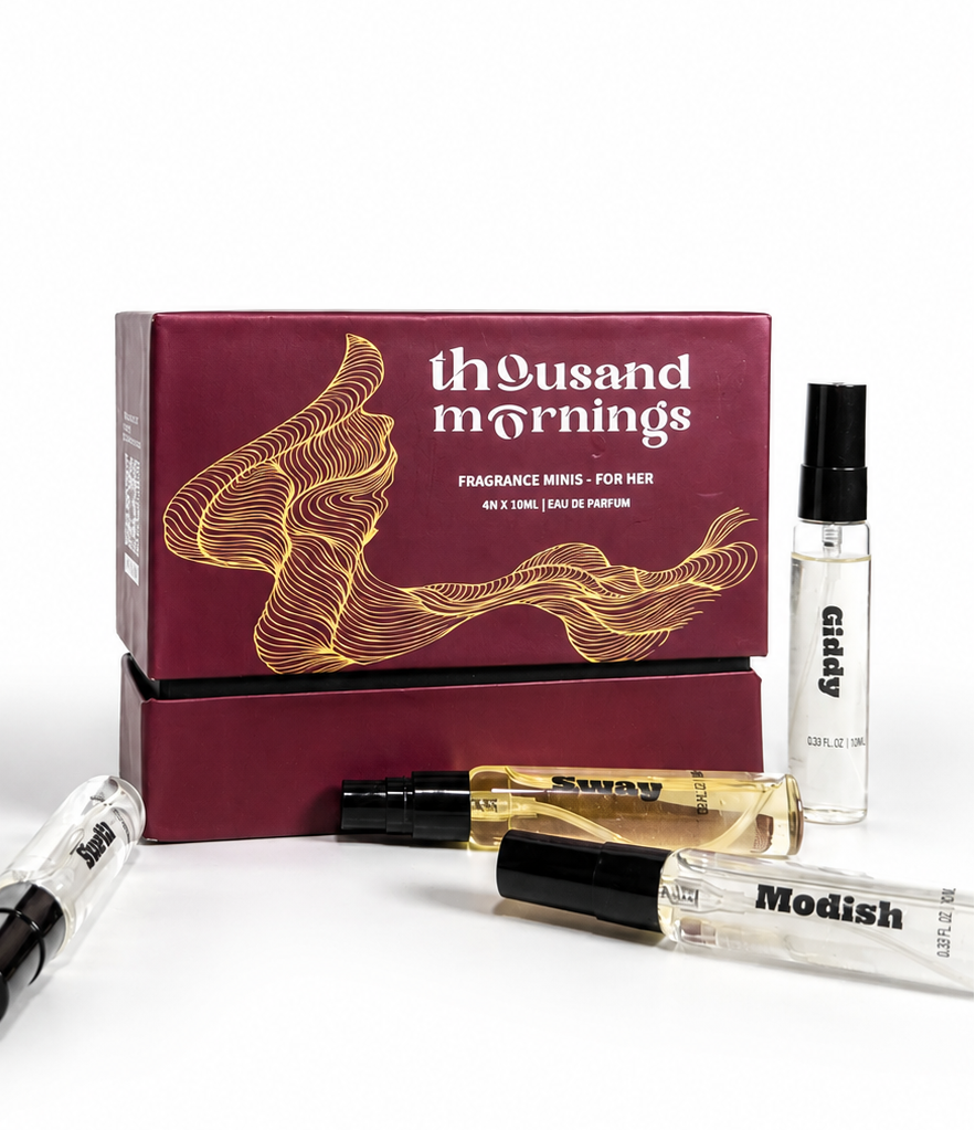 A fragrance oil set featuring a dark burgundy box with gold line art and white text reading 'thousand mornings' and 'FRAGRANCE OIL FOR MEN'. In front of the box are two clear glass perfume oil vials with black caps, one labeled 'Sakura' and the other 'Modish', alongside a small clear vial labeled 'caddy' on the right, all on a clean white background.