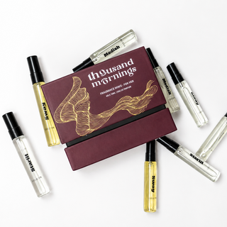 A perfume gift set featuring a dark burgundy box labeled 'thousand mornings' with gold line art, surrounded by several clear glass rollerball vials with black caps and amber liquid, arranged on a white background.