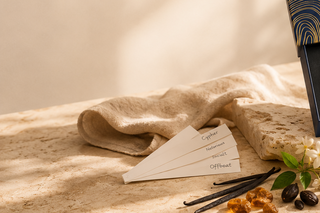 A folded beige linen towel and a stack of cream color sample cards labeled Cypher, Neutral, Warm, and Offbeat, styled on a stone surface with vanilla beans, cocoa nibs, and a small white flower.