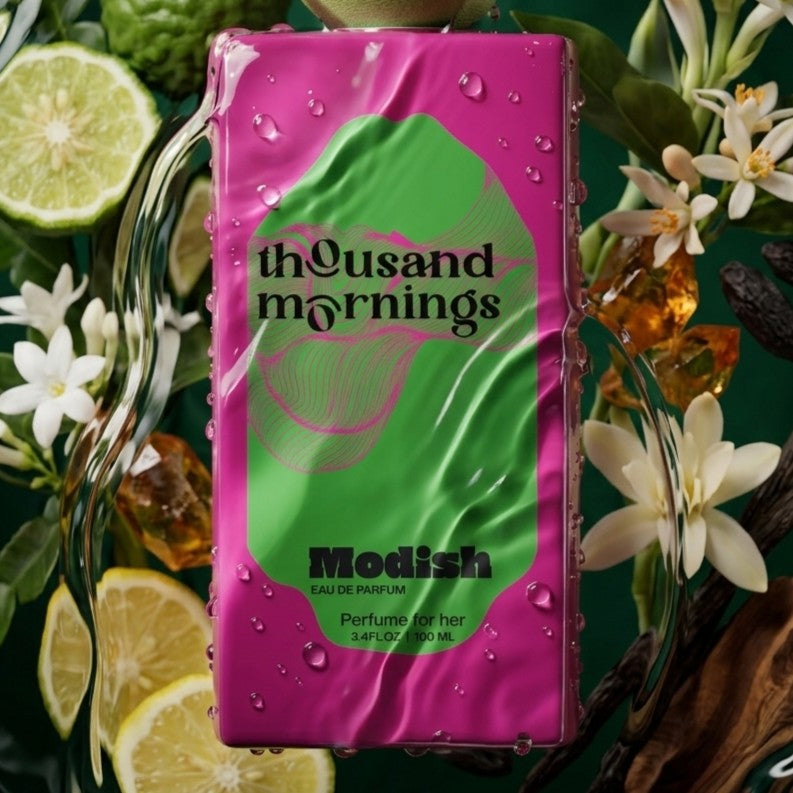 A rectangular perfume bottle labeled 'thousand mornings' and 'Modish' with a bright pink and green label, surrounded by citrus slices, white flowers, and amber droplets on a dark green background.